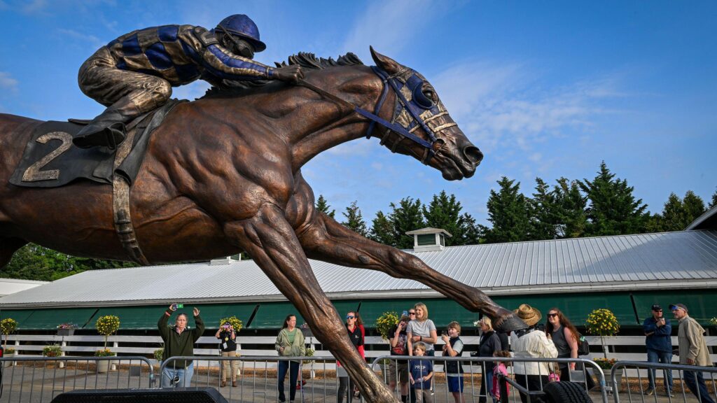 Bronze statue of Secretariat and jockey racing forward at Pimlico Race Course, with visitors below under a blue sky.