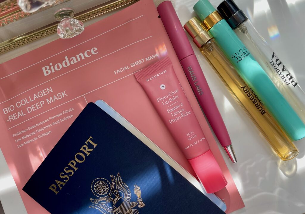 Passport beside a Biodance sheet mask, Naturium lip balm, Haus Labs lip liner, and travel-size fragrances arranged on a mirrored tray.